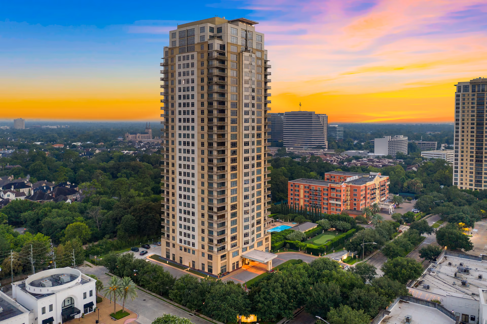 Aerial photograph of Montebello at sunset: a cream-colored high-rise with recessed corner balconies and a stepped crown stands at the center of the frame, its west face lit warm by the low sun, above a lit covered entrance at street level. In the grounds to its right are a swimming pool, a small putting green and clipped hedges. Low houses and heavy tree cover fill the left half, a red-brick mid-rise stands to the right, a second cream high-rise sits at the right edge, and mid-rise office blocks stand on the horizon under a sky graded from blue at the top to orange and pink along the skyline.